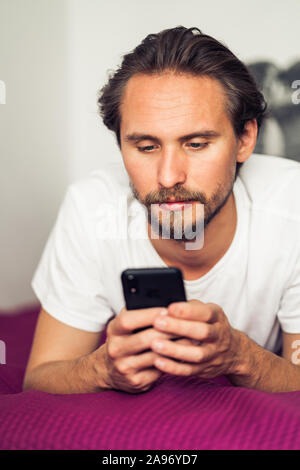 Foto di un piuttosto grave giovane uomo bello interagire con il suo dispositivo mobile mentre giaceva a letto Foto Stock