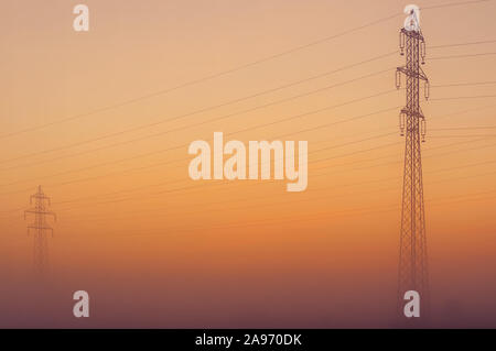 Torri di trasmissione uscente dalla nebbia con orange twilight sky in background. L'elettricità, la potenza di trasmissione e distribuzione di energia elettrica Foto Stock