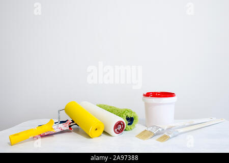 Un secchio di vernice, una serie di rulli colorati e spazzole su un tavolo bianco contro un bianco parete dipinta. Foto Stock