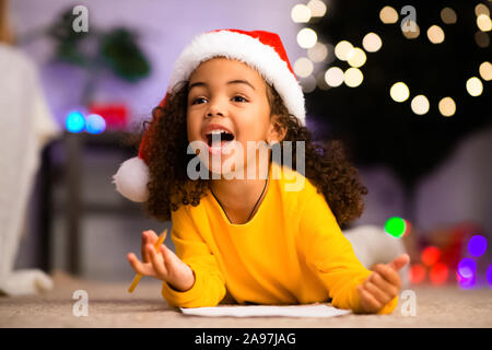 Piccolo felice nero ragazza di disegno la lista dei desideri per Babbo Natale, giacente sul piano a casa Foto Stock