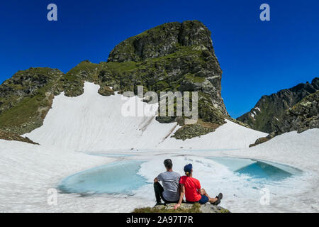 Un giovane godendo la vista di un lago alpino congelato. Il ghiaccio brilla in molte diverse sfumature di blu. Il lago è circondato da alte montagne. Assenza di neve sul Foto Stock