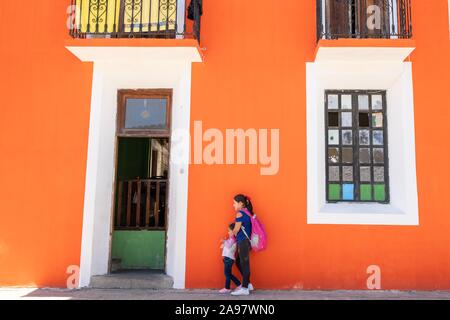 Un messicano di donna e di sua figlia di attendere per un autobus di fronte a un dipinto luminosamente edificio arancione in Xico, Veracruz, Messico. Foto Stock