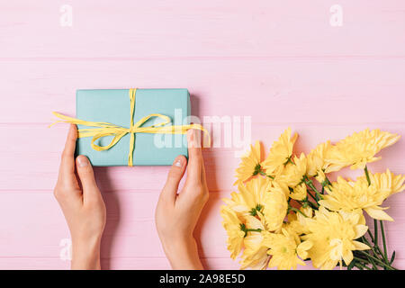 Mani femminili azienda confezione regalo accanto al bouquet di fiori freschi su sfondo rosa, piatto laici. Foto Stock