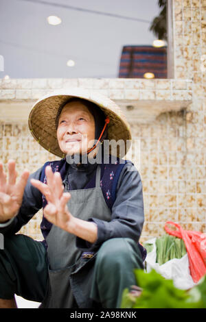 Sorridente donna senior accovacciato in un luogo di mercato indossando un cappello. Foto Stock