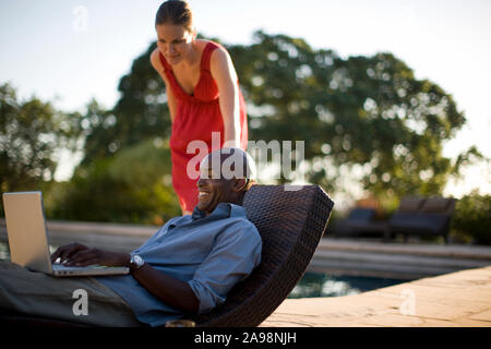 Metà di uomo adulto su un portatile con la moglie a bordo della piscina al tramonto. Foto Stock