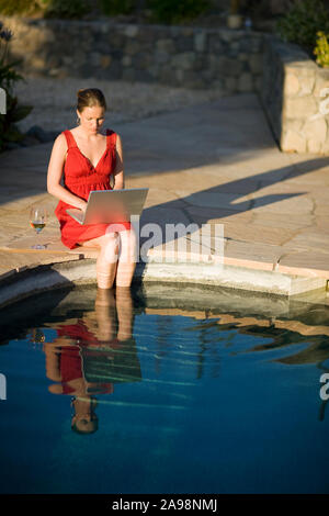 Metà donna adulta Dondolando i suoi piedi in piscina mentre su un laptop. Foto Stock