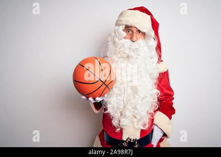 Senior uomo che indossa Santa Claus costume holding basket ball isolato su sfondo bianco paura in stato di shock con una sorpresa faccia paura ed entusiasti Foto Stock