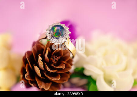 Opale anello solitario circondato da diamanti in una forma rotonda con fascia in oro su un cono di pino con fiori e un sfondo rosa Foto Stock