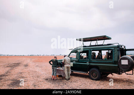 Giu 21, 2011 Serengeti Tanzania - turisti e tavolo da picnic accanto a AUTO Jeep Safari carrello in golden campo in erba della savana del Serengeti foresta in tanza Foto Stock