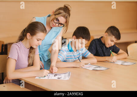 Insegnante di scuola aiuta i ragazzi a scrivere test in aula. Istruzione, scuola elementare, apprendimento e concetto di persone Foto Stock