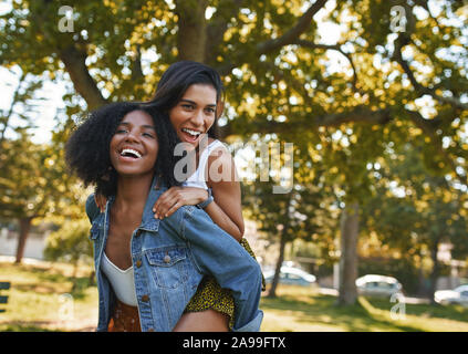 Due femmina multietnica migliori amici divertendosi e giocare nel parco - due amici dando loro una piggy back e ridere all'aperto nel parco Foto Stock