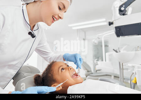 Dentista femmina in camice bianco e guanti di gomma il trattamento di denti della piccola ragazza africana. Paziente seduto in poltrona odontoiatrica con bocca aperta. Medico azienda strumenti dentali e guardando i denti. Foto Stock