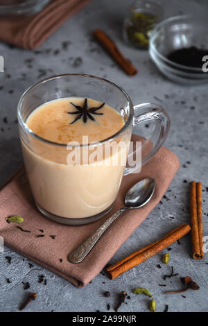 Masala chai. Indian Black Tea con spezie e il latte in un bicchiere di vetro su uno sfondo grigio. Foto verticale, close-up. Foto Stock