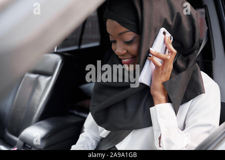 Messa a fuoco selettiva della bella ragazza islamica vestita di nero hijab seduto in automobile grande e parla al telefono. Donna musulmana guardando verso il basso e ridere mentre si guida. Concetto di automobile e comfort. Foto Stock