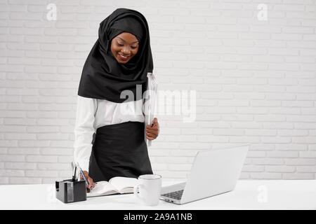Vista ritagliata della giovane donna africana vestita di nero hijab in piedi in ufficio e lavoro. Attraente femminile musulmano iscritto nel notebook e uso del computer. Concetto di multi etnico e società. Foto Stock