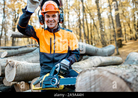 Ritratto di un allegro professional lumberjack in indumenti da lavoro protettiva in piedi con una motosega su una pila di registri nella foresta Foto Stock