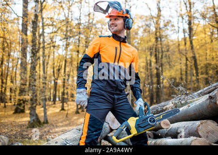 Ritratto di un bel professional lumberjack in indumenti da lavoro protettiva con un chainsaw vicino i tronchi di legno nella foresta Foto Stock