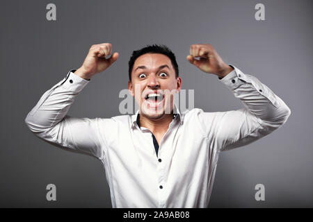 Angry Young business man looking aggressiva con gli occhi e agitando le mani con i pugni sopra la testa su sfondo grigio. Closeup emozione ritratto Foto Stock