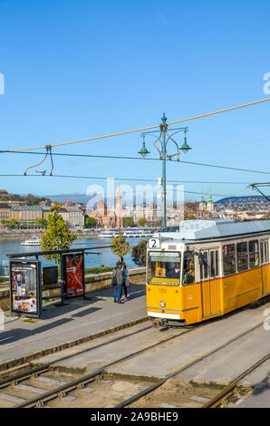 Budapest, Ungheria - Novembre 6, 2019: tram giallo in corrispondenza di una stazione di pubblico. Il fiume Danubio e la città vecchia in background. Persone presso la stazione del tram. Giornata di sole. Foto verticale. Foto Stock