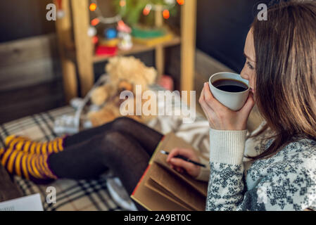 Una giovane donna in un maglione è scrivere una lista dei desideri in un notebook con una tazza di caffè la sera nella calda atmosfera del Natale. Accogliente Anno Nuovo Foto Stock