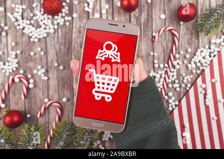 Concetto per la stagione di Natale regalo online shopping con mano azienda telefono cellulare con il bianco carrello segno su sfondo rosso nella parte anteriore del legno de Foto Stock