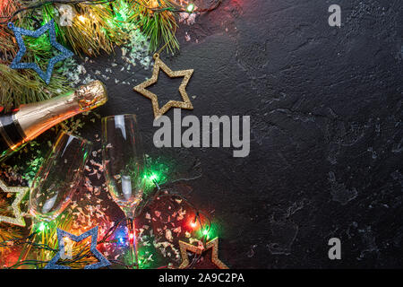 Immagine di rami di abete, decorazioni di Natale blu e oro per giocattoli, bruciando le ghirlande, bottiglie di champagne, 2 bicchiere di vino su sfondo nero, posto per Foto Stock