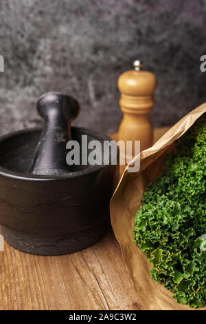 Ingredienti per il cavolo verde chip: Kale in sacchetto di carta, nero mortaio e pestello in legno mulino di pepe su una superficie di legno scuro dello sfondo testurizzata Foto Stock