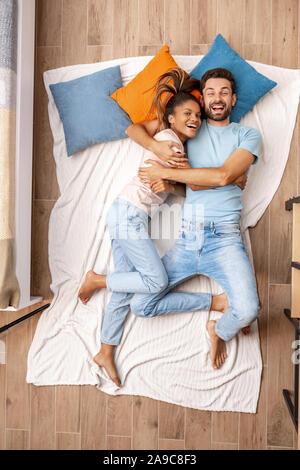 Razza mista giovane. Giovane uomo e donna sdraiata sul letto vista dall'alto abbracciando ridendo allegro Foto Stock