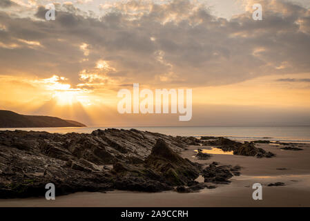 alba con fasci di luce dorata, rocce in primo piano con sabbia, con cielo giallo e nuvole Foto Stock