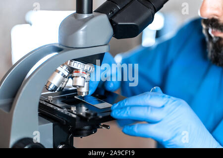 Maschio assistente di laboratorio esaminando campioni di biomateriale in un microscopio. Cllose fino a mani in blu di guanti di gomma regolare il microscopio. Foto Stock