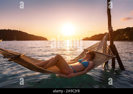 Donna di relax presso la spiaggia in un'amaca sopra l'acqua di mare, sole calda serata estiva a vacanza vacanze hotel, momento felice al tramonto
