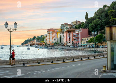 Un uomo cammina sul marciapiede accanto al porto marina a Villefranche Sur Mer, Francia, come il sole che tramonta dietro gli edifici colorati. Foto Stock