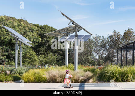 I bambini giocano e ride scooter vicino polo regolabile montato su pannelli solari in un giardino e un parco pubblico a sfere riserva di testa a Sydney in Australia Foto Stock