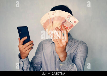 Giovane uomo detiene il denaro in mano e si preme lo schermo dello smartphone. Guadagna denaro attraverso il mobile app. Business via smartphone. Foto Stock