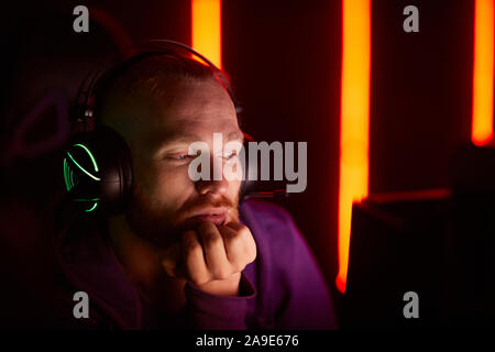 Giovane uomo barbuto indossare cuffie per giochi con retroilluminazione guardando il monitor del computer pensively Foto Stock