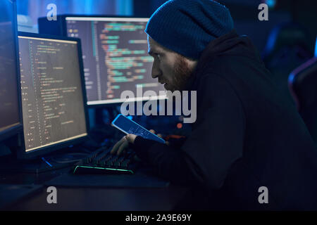 Hacker in abbigliamento scuro seduti nella parte anteriore del monitor per computer e utilizzando il suo telefono cellulare di introdursi nel sistema Foto Stock