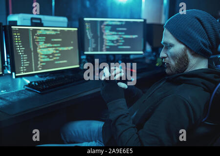 Barbuto giovani hacker nel cappello e maglietta felpa con cappuccio seduti nella parte anteriore del monitor per computer con software e utilizzando il telefono cellulare nella camera oscura Foto Stock