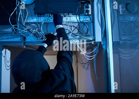 Vista posteriore dell'uomo in felpa con cappuccio tirando fuori i fili e spegnendo il computer ha intenzione di rendere il crimine Foto Stock