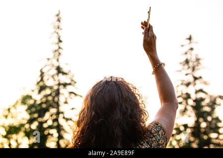 Una donna caucasica è visto da dietro mentre lei solleva un pezzo di corteccia di albero verso il cielo al tramonto, offuscata alberi in background Foto Stock