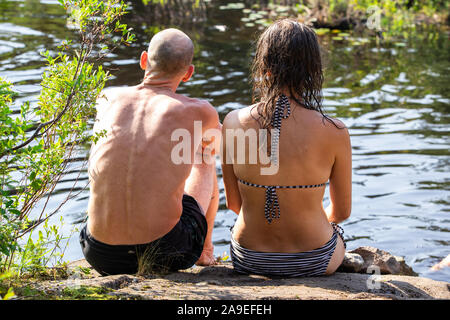Giovane uomo e donna vestito in costume da bagno sono seduto su una grande roccia sul lato di un lago. Immagine presa dal retro all'aperto in estate Foto Stock