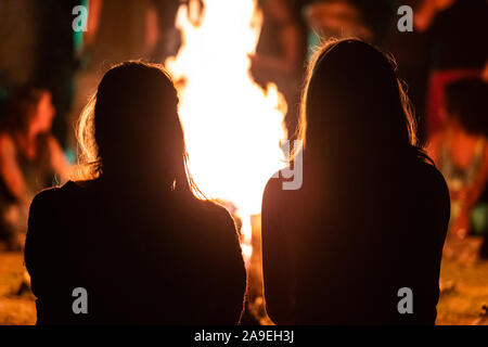 Silhouette di due ragazze seduti davanti al fuoco come si guarda la notte spettacolo musicale, visto da dietro durante la notte oscura falò, sfondo sfocato Foto Stock