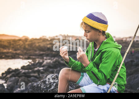 Giovane pescatore con la pesca con sistemi di poli la linea seduta sulle rocce vicino al porto. Sailor ragazzo con giacca cerata godendo di estate la vigilia del tramonto Foto Stock