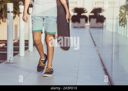 Bambino alla moda a camminare in una città moderna di contesto. Ragazzo con lo skateboard in mano cammina da sola. Vista ravvicinata del giovane maschio gambe camminando Foto Stock