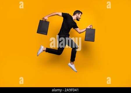 Lunghezza completa di foto bello ragazzo millenario jumping alta acceso shopping mall comprare vestiti nuovi bagagli da stiva indossare nero t-shirt pantaloni giallo isolato Foto Stock