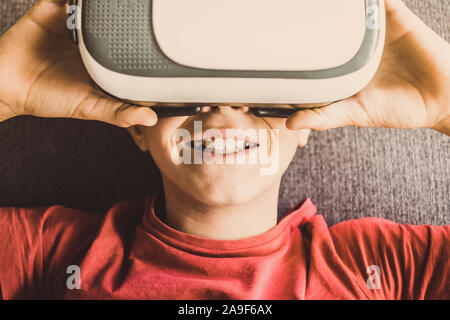 Giovane uomo divertendosi con la realtà virtuale tecnologia occhiali sdraiato sul divano di casa. Stupito sorriso del ragazzo che indossa le cuffie vr. Tech, lo stile di vita moderno Foto Stock