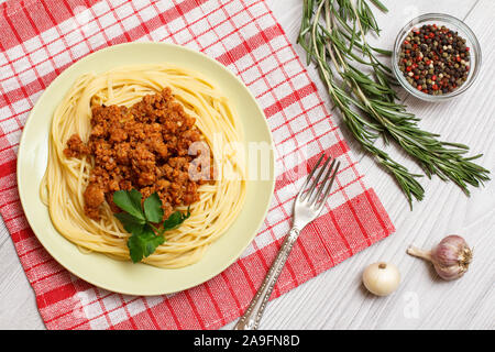 La piastra di porcellana con gli spaghetti e il ragù alla bolognese, forcella, rosmarino e ciotola di vetro con pepe della Giamaica peperoni su un tovagliolo da cucina. Vista dall'alto. Foto Stock