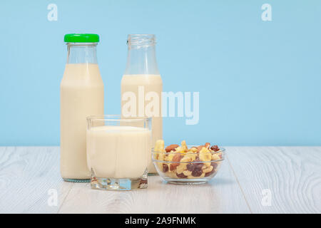 Chiuso, aperto bottiglie e contenitori di vetro di latte fresco, ciotola di vetro con muesli su sfondo blu. Foto Stock