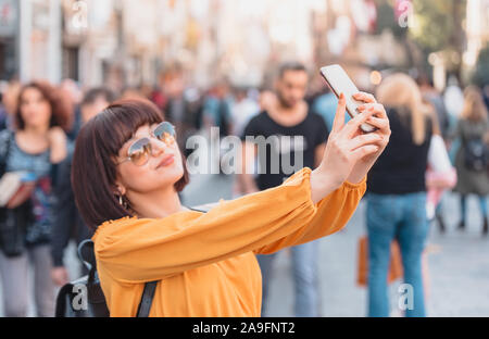 Giovane e bella ragazza in abiti alla moda prende selfie ad una strada affollata strada.Selfie concetto. Foto Stock