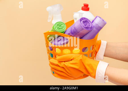 Donna con le mani in mano in gomma guanti protettivi azienda cesto arancione con sacchi della spazzatura, bottiglie di vetro e tile cleaner, spugna su sfondo beige. Lavare Foto Stock