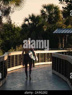 Giovane biondo surfer tenendo la sua tavola da surf cammina per il New Smyrna Dune Boardwalk per la spiaggia di sunrise. Foto Stock